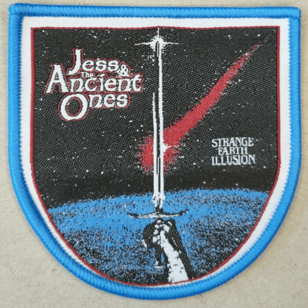 Jess and the Ancient Ones sleeve patch Strange Earth Illusion live pic on grey background. Shape of patch is round bottom and straight up side. Pic in patch is hand holding sword up with texts Jess & the Ancient Ones and Strange Earth Illusion.